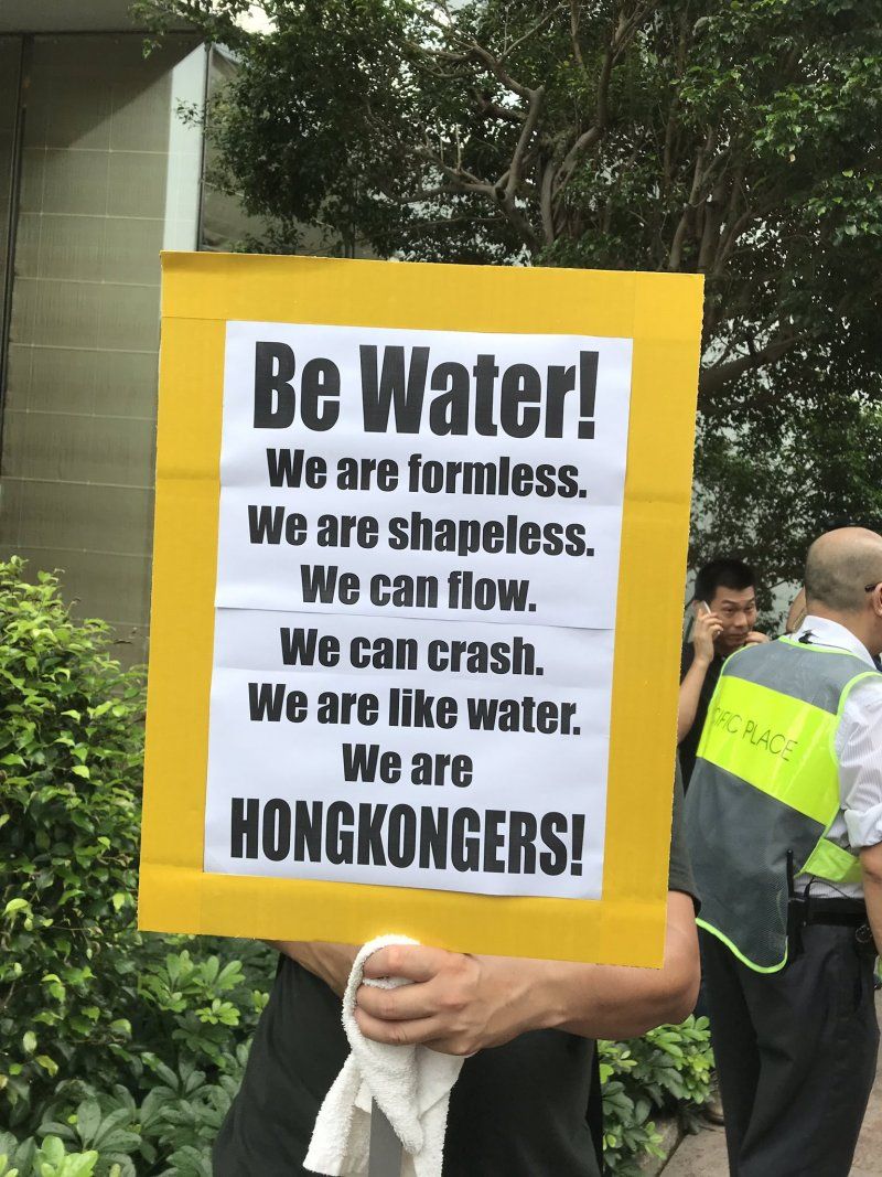 Photograph of a protester holding a yellow sign reading "Be Water! We are formless. We are shapeless. We can flow. We can crash. We are like water. We are HONGKONGERS!"